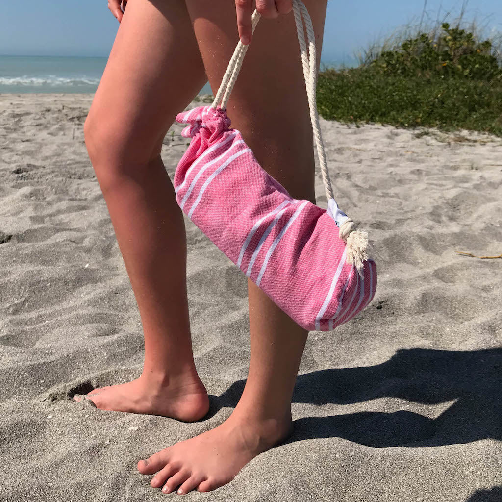 girl standing on sand holding pink perim hammam towel in carry pouch